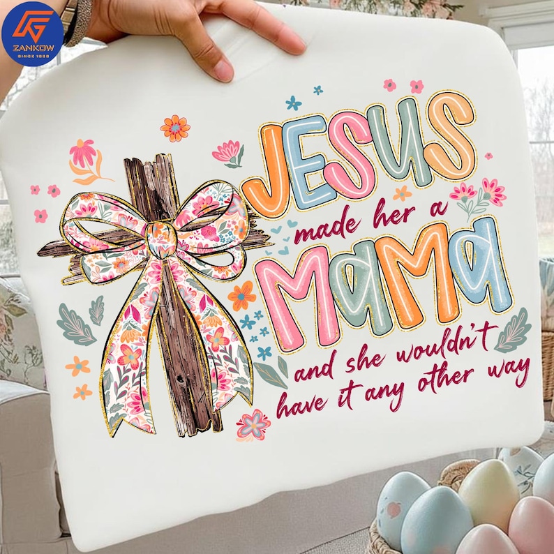 May include: White t-shirt with the text "JESUS made her a MAMA and she wouldn't have it any other way." The shirt features a wooden cross with a floral bow and colourful floral accents.