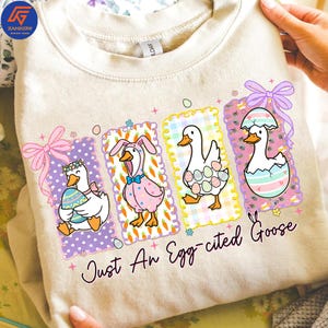 May include: A beige sweatshirt featuring four cartoon geese in Easter-themed outfits. The geese are in colorful frames with carrots and eggs. The text "Just An Egg-cited Goose" is printed below the geese.
