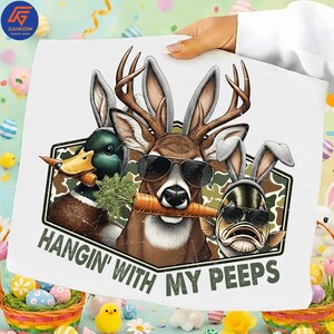 May include: White graphic tee featuring a deer, duck, and fish wearing sunglasses and bunny ears, each holding a carrot. The text "HANGIN' WITH MY PEEPS" is printed below the image. The design has a camouflage background.