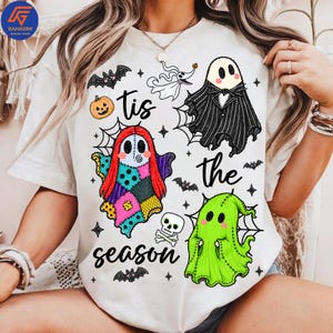 May include: A white t-shirt featuring a graphic print with the text 'tis the season' and various Halloween-themed characters including a ghost in a black suit, a patchwork ghost, a green ghost, a small white dog ghost, a pumpkin, a skull, and bats.