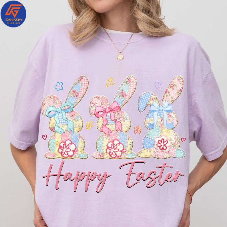 May include: A light purple t-shirt featuring three appliqued Easter bunnies with floral and patchwork patterns. Each bunny has a different colored bow and a flower at the bottom. The words "Happy Easter" are written in pink script below the bunnies.