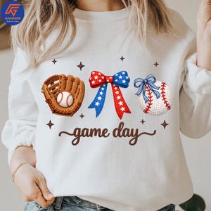 May include: White sweatshirt with a baseball theme. Features a baseball glove with a baseball, a red, white, and blue bow, a baseball with red stitching, and the words "game day". Decorative stars are also included.