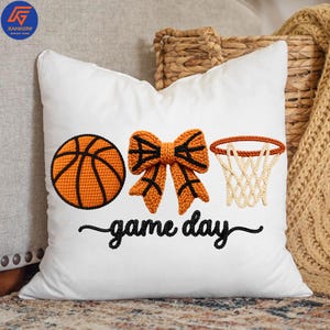 May include: White square pillow with an embroidered basketball, bow, and hoop design. The basketball is orange with black lines, the bow is orange with black accents, and the hoop has a white net. The words "game day" are embroidered in black.