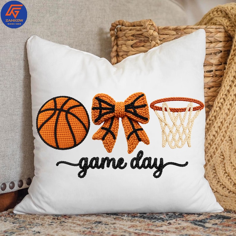 May include: White square pillow with an embroidered basketball, bow, and hoop design. The basketball is orange with black lines, the bow is orange with black accents, and the hoop has a white net. The words "game day" are embroidered in black.
