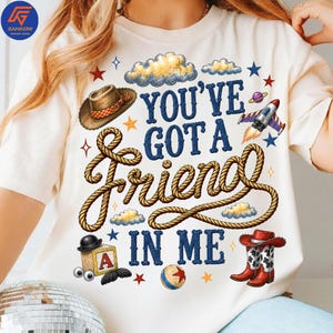 May include: A cream-colored t-shirt featuring a graphic print with the text "YOU'VE GOT A FRIEND IN ME" in blue letters. The design includes a brown cowboy hat, a coiled rope, a rocket ship, a wooden block with the letter A, and cowboy boots.