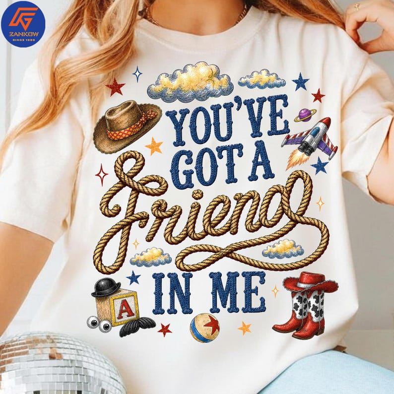 May include: A cream-colored t-shirt featuring a graphic print with the text "YOU'VE GOT A FRIEND IN ME" in blue letters. The design includes a brown cowboy hat, a coiled rope, a rocket ship, a wooden block with the letter A, and cowboy boots.