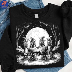 May include: Black sweatshirt featuring a graphic of four witches holding hands in front of a full moon. The witches are wearing pointed hats and long dresses. The text "GIRLS WILL BE GIRLS" is printed below the image.