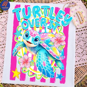 May include: White t-shirt featuring a colorful cartoon turtle with the text "TURTLES OVER IT" in turquoise. The turtle has a blue body, pink and yellow shell, and is surrounded by flowers and stars. The design is set against a pink and white striped background.