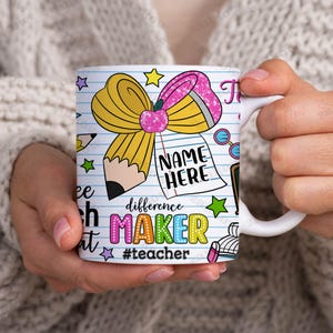 May include: White ceramic mug with a colorful design featuring a yellow pencil bow, a notepad that says "NAME HERE", and the words "difference MAKER #teacher". The mug is held by a person.