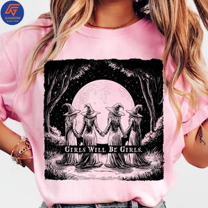 May include: Pink t-shirt featuring a black and white graphic of four witches holding hands in front of a full moon. The text "GIRLS WILL BE GIRLS" is printed below the image. The shirt has short sleeves.