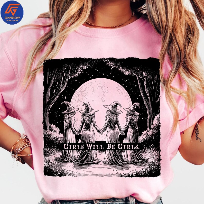 May include: Pink t-shirt featuring a black and white graphic of four witches holding hands in front of a full moon. The text "GIRLS WILL BE GIRLS" is printed below the image. The shirt has short sleeves.