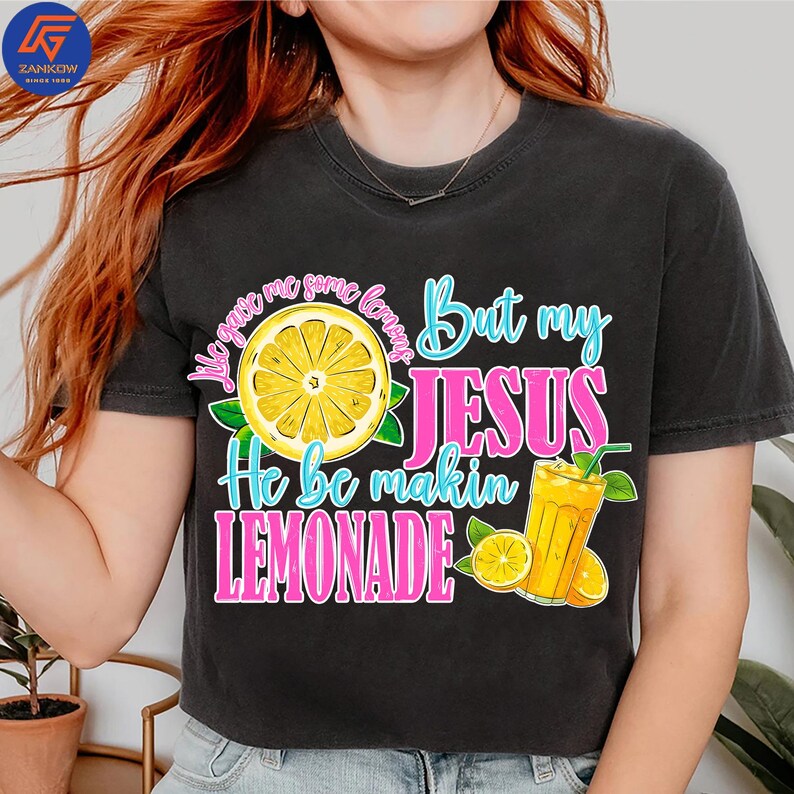 May include: Black t-shirt with a colorful graphic design. The design features a yellow lemon slice, a glass of lemonade, and the text "He be makin LEMONADE". The text is in pink and blue.