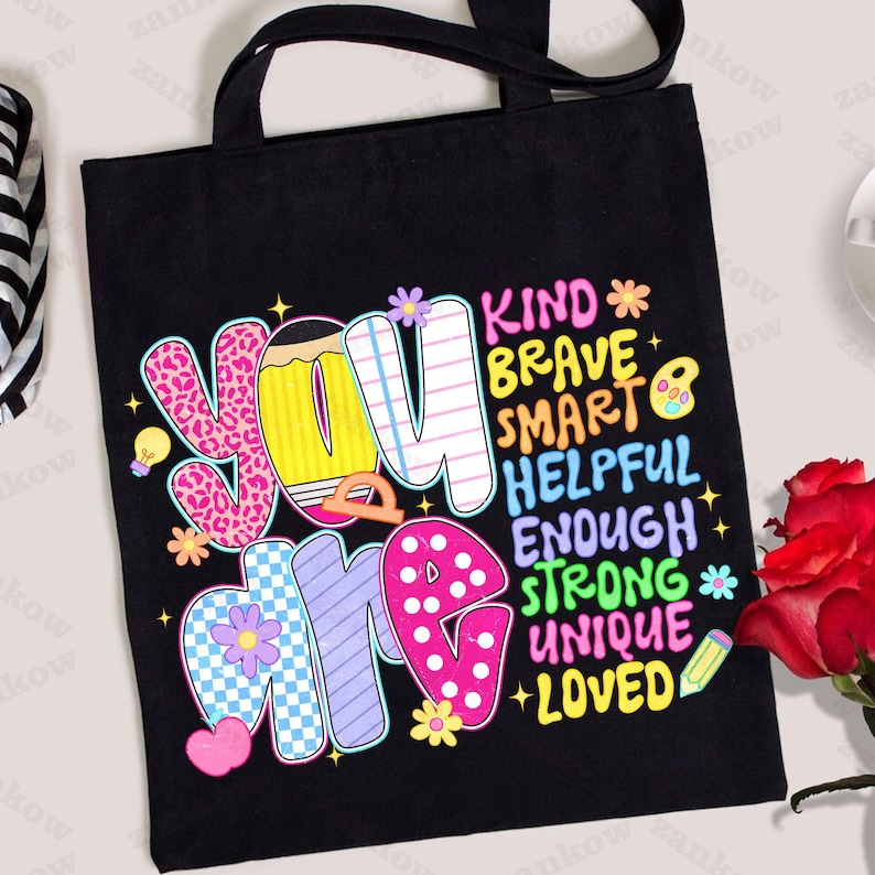 May include: Black tote bag with the words "YOU ARE" in colorful, patterned letters. The words "KIND, BRAVE, SMART, HELPFUL, ENOUGH, STRONG, UNIQUE, LOVED" are also printed on the bag. The bag has black handles.
