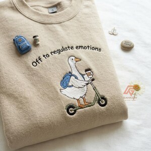 Op de afbeelding: Een beige sweater met ronde hals en een geborduurde afbeelding van een witte gans op een groene step, met een blauwe rugzak en een koffiebeker in de hand. De tekst "Off to regulate emotions" is boven de gans geborduurd.