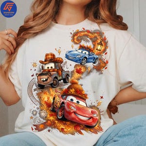 Puede incluir: Una camiseta blanca con un estampado gráfico de tres coches animados de la película Cars. El diseño incluye una grúa marrón con un sombrero de cuadros, un coche deportivo azul y un coche de carreras rojo con un gorro de punto, entre hojas de otoño.