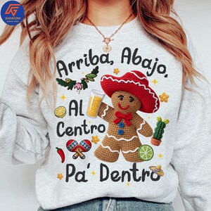 May include: A light gray sweatshirt with the text "Arriba, Abajo, Al Centro, Pa' Dentro". The design includes a crochet gingerbread man wearing a red sombrero, holding a drink, and surrounded by festive Mexican-themed graphics.