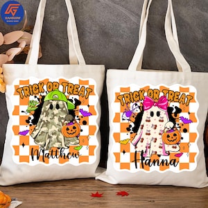 May include: Two white canvas tote bags with Halloween-themed designs. One features a ghost in a camo outfit, a green hat, and a pumpkin, with the name "Matthew." The other has a ghost with a pink bow, holding a pumpkin, with the name "Hanna."