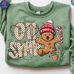 May include: A sage green sweatshirt featuring a gingerbread man with a red knit cap and candy cane. The phrase "OH SNAP" is displayed in a checkered pattern with colorful Christmas lights. The gingerbread man has a broken leg and is surrounded by cookie crumbs.