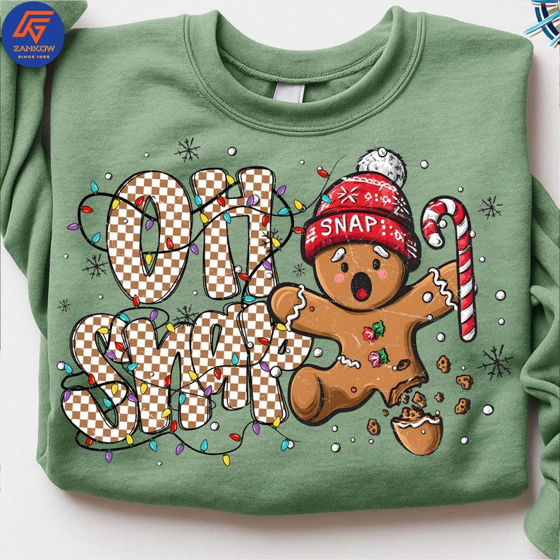 May include: A sage green sweatshirt featuring a gingerbread man with a red knit cap and candy cane. The phrase "OH SNAP" is displayed in a checkered pattern with colorful Christmas lights. The gingerbread man has a broken leg and is surrounded by cookie crumbs.