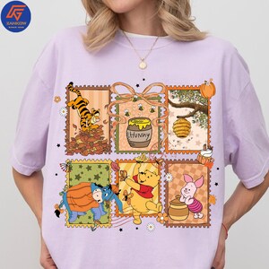 May include: A lilac t-shirt with a colourful graphic of Winnie the Pooh characters in postage stamp designs. The design includes Tigger, Winnie the Pooh, Piglet, Eeyore, a honey pot, and a beehive.