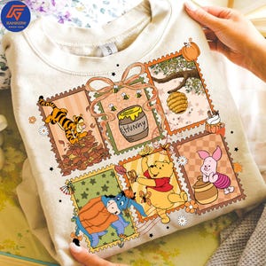 May include: Cream-coloured sweatshirt with a colourful graphic of Winnie the Pooh characters in postage stamp designs. The design includes Tigger, Winnie the Pooh, Piglet, Eeyore, a honey pot, and a beehive.
