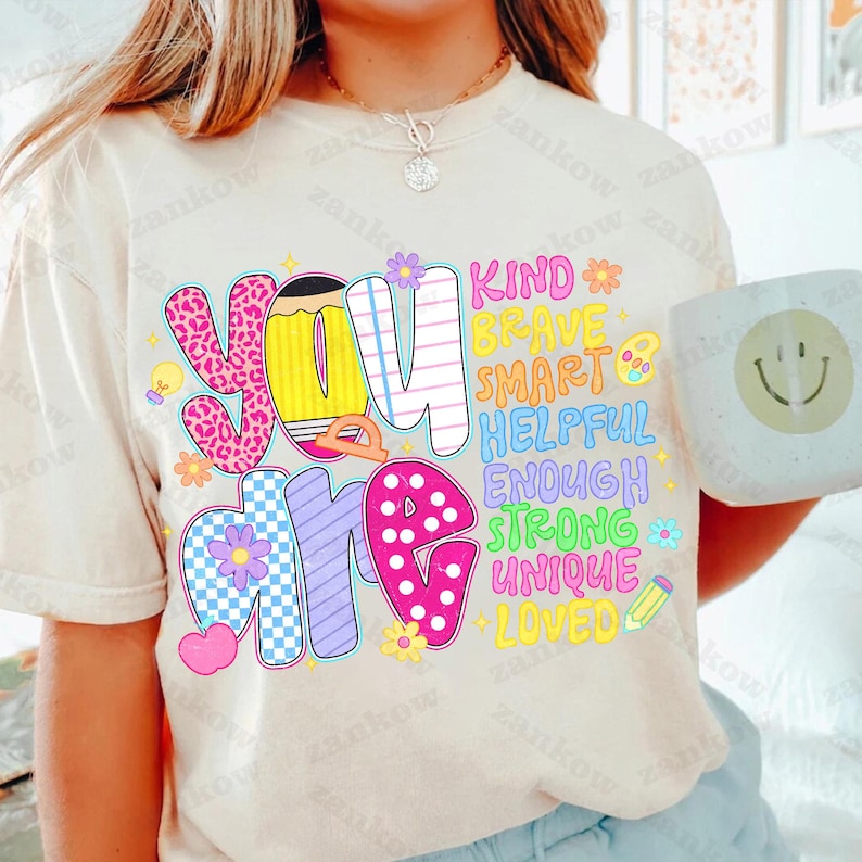 May include: Cream-colored t-shirt with the words "You Are" in colorful, playful lettering. The words "Kind, Brave, Smart, Helpful, Enough, Strong, Unique, Loved" are also displayed. A white mug with a smiley face is held in the image.