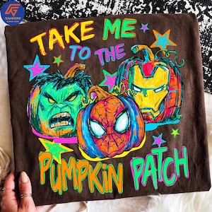 May include: A brown t-shirt featuring a graphic print of three pumpkins carved to resemble superhero faces, including the Hulk, Spider-Man, and Iron Man. The text reads TAKE ME TO THE PUMPKIN PATCH in bright yellow, pink, and green lettering.