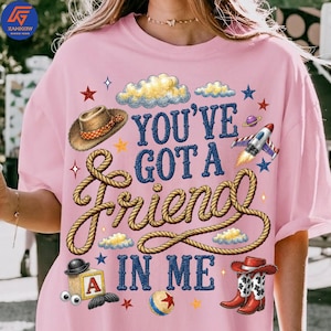 May include: A light pink t-shirt featuring a graphic print with the text "YOU'VE GOT A FRIEND IN ME" in blue. The design includes a brown cowboy hat, a rope, a rocket ship, a toy block with the letter A, a ball, and red cowboy boots.