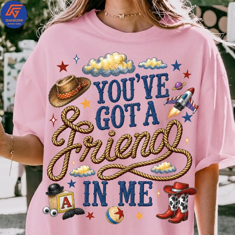 May include: A light pink t-shirt featuring a graphic print with the text "YOU'VE GOT A FRIEND IN ME" in blue. The design includes a brown cowboy hat, a rope, a rocket ship, a toy block with the letter A, a ball, and red cowboy boots.