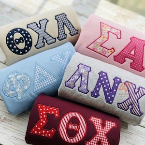 May include: Five folded sweatshirts in cream, light blue, pink, grey, and maroon, each featuring large Greek letters appliqued in various patterns including stars, stripes, gingham, polka dots, and floral prints with stitched borders.