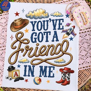 May include: A white t-shirt featuring the dark blue text YOU'VE GOT A FRIEND IN ME. The design includes a brown cowboy hat, a coiled rope, a rocket ship, a wooden block with the letter A, a ball, and red cowboy boots, surrounded by stars and clouds.