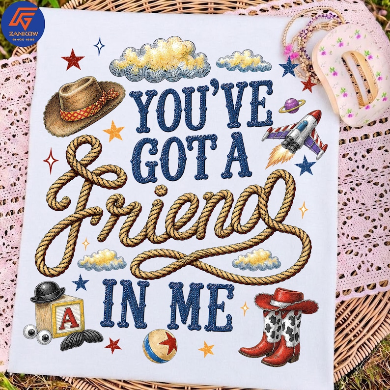 May include: A white t-shirt featuring the dark blue text YOU'VE GOT A FRIEND IN ME. The design includes a brown cowboy hat, a coiled rope, a rocket ship, a wooden block with the letter A, a ball, and red cowboy boots, surrounded by stars and clouds.