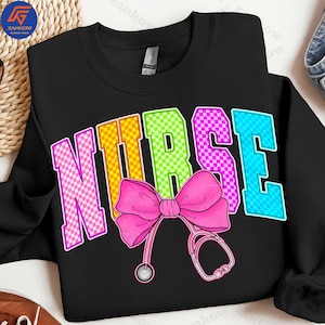 K&ouml;nnte beinhalten: Schwarzes Sweatshirt mit dem Wort "NURSE" in bunten, karierten Buchstaben. Eine rosa Schleife und eine Stethoskop-Grafik befinden sich darunter. Das Sweatshirt hat einen Rundhalsausschnitt.
