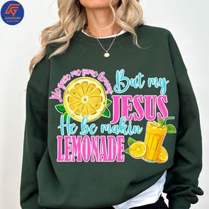 May include: Dark green sweatshirt with the text "They gave me some lemons But my JESUS He be makin LEMONADE" in pink and blue. The design includes a lemon slice and a glass of lemonade.