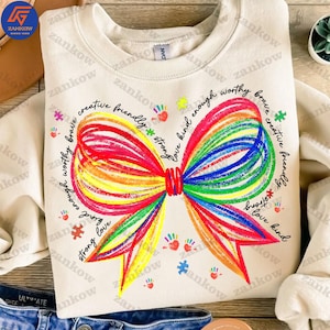 May include: A beige sweatshirt with a rainbow-colored bow tie design. The bow tie is made of colorful lines and has the words "love kind enough worthy brave creative friendly strong" written around it.
