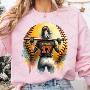 K&ouml;nnte beinhalten: Rosafarbener Pullover mit einem Aquarell-Design einer Softballspielerin, die ein schwarzes Trikot mit dem Namen "Emma" und der Nummer "17" auf dem R&uuml;cken tr&auml;gt. Die Spielerin steht vor einem gelben Softball.