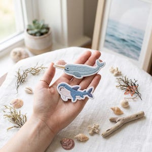 Op de afbeelding: Twee geborduurde stoffen patches met een lichtblauwe narwal met een gele slagtand en een blauw-witte haai met een glimlachende uitdrukking. Beide patches van zeedieren hebben gedetailleerd stikwerk en volgen de contouren van de figuren.