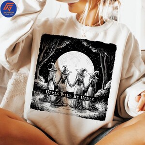 May include: White sweatshirt featuring a black and white graphic of four witches holding hands in a forest under a full moon. The text "GIRLS WILL BE GIRLS" is printed below the image.