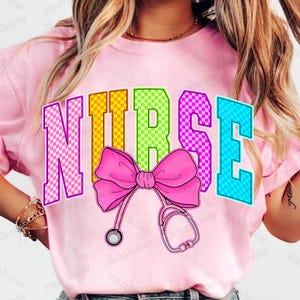 K&ouml;nnte beinhalten: Hellrosa T-Shirt mit dem Wort "NURSE" in bunten, karierten Buchstaben. Eine rosa Schleife und eine Stethoskop-Grafik sind unter dem Wort zentriert. Das Shirt hat eine hellrosa Farbe.