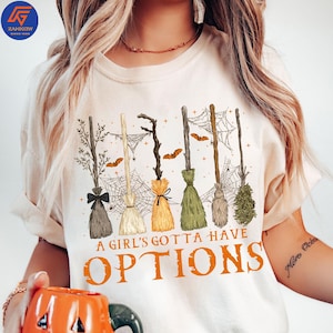 以下が含まれることがあります： ハロウィーンをテーマにしたデザインのクリーム色のTシャツ。グラフィックには、クモの巣とコウモリが付いたいくつかの魔女のほうきと、オレンジ色のテキスト「A Girl's Gotta Have Options」が含まれています。ほうきはさまざまな色の毛を持っています。