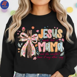 May include: Black sweatshirt featuring the text "JESUS made her a MAMA and she wouldn't have it any other way." The design includes a wooden cross with a floral bow and colourful flowers.