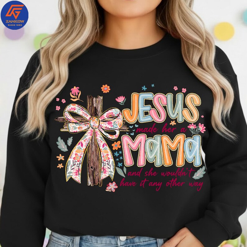 May include: Black sweatshirt featuring the text "JESUS made her a MAMA and she wouldn't have it any other way." The design includes a wooden cross with a floral bow and colourful flowers.
