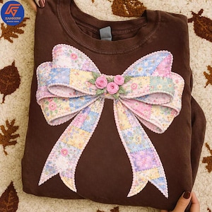 May include: A brown sweatshirt featuring a large, decorative bow. The bow is made of patchwork fabric in pastel colors, with floral patterns and a scalloped edge. Three small pink roses are at the bow's center. The sweatshirt has a crew neck.
