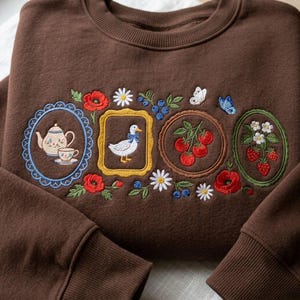 May include: A dark brown crewneck sweatshirt featuring intricate embroidery of four framed scenes. The designs include a teapot and teacup, a white duck with a blue bow, red tomatoes, and strawberries, all surrounded by small flowers and butterflies.
