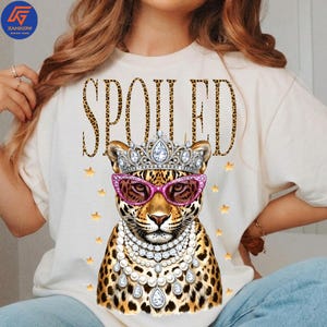 May include: Cream-colored t-shirt featuring a leopard illustration wearing a jeweled crown, pink sunglasses, and a pearl necklace. The word "SPOILED" is printed above the leopard in a leopard print font. Gold stars are scattered around the leopard.