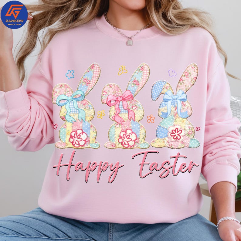 May include: A light pink sweatshirt featuring three patchwork bunny rabbits with floral and gingham patterns. Each bunny has a bow and a flower. The words "Happy Easter" are written in pink script below the bunnies.