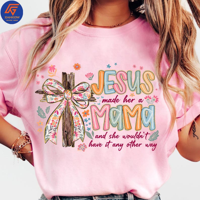 May include: Pale pink t-shirt featuring the text "JESUS made her a MAMA and she wouldn't have it any other way." The design includes a wooden cross with a floral bow and colorful floral accents. The shirt has short sleeves.