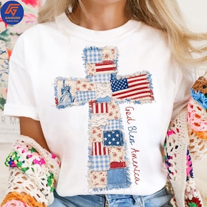 May include: White t-shirt featuring a cross design made of patchwork fabric. The cross incorporates elements of the American flag, the Statue of Liberty, and floral patterns. The words "God Bless America" are written in red.