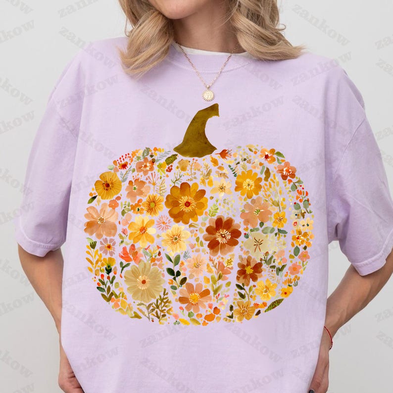 May include: A light purple t-shirt featuring a large pumpkin design. The pumpkin is filled with a variety of colorful flowers in shades of orange, yellow, and brown, with a gold stem. The shirt is a comfortable, casual style.