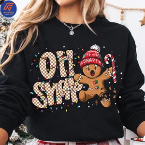 May include: Black sweatshirt with a festive design featuring the words "Oh Snap" in a checkered pattern, surrounded by colorful lights. A gingerbread man with a red hat and candy cane completes the holiday theme.